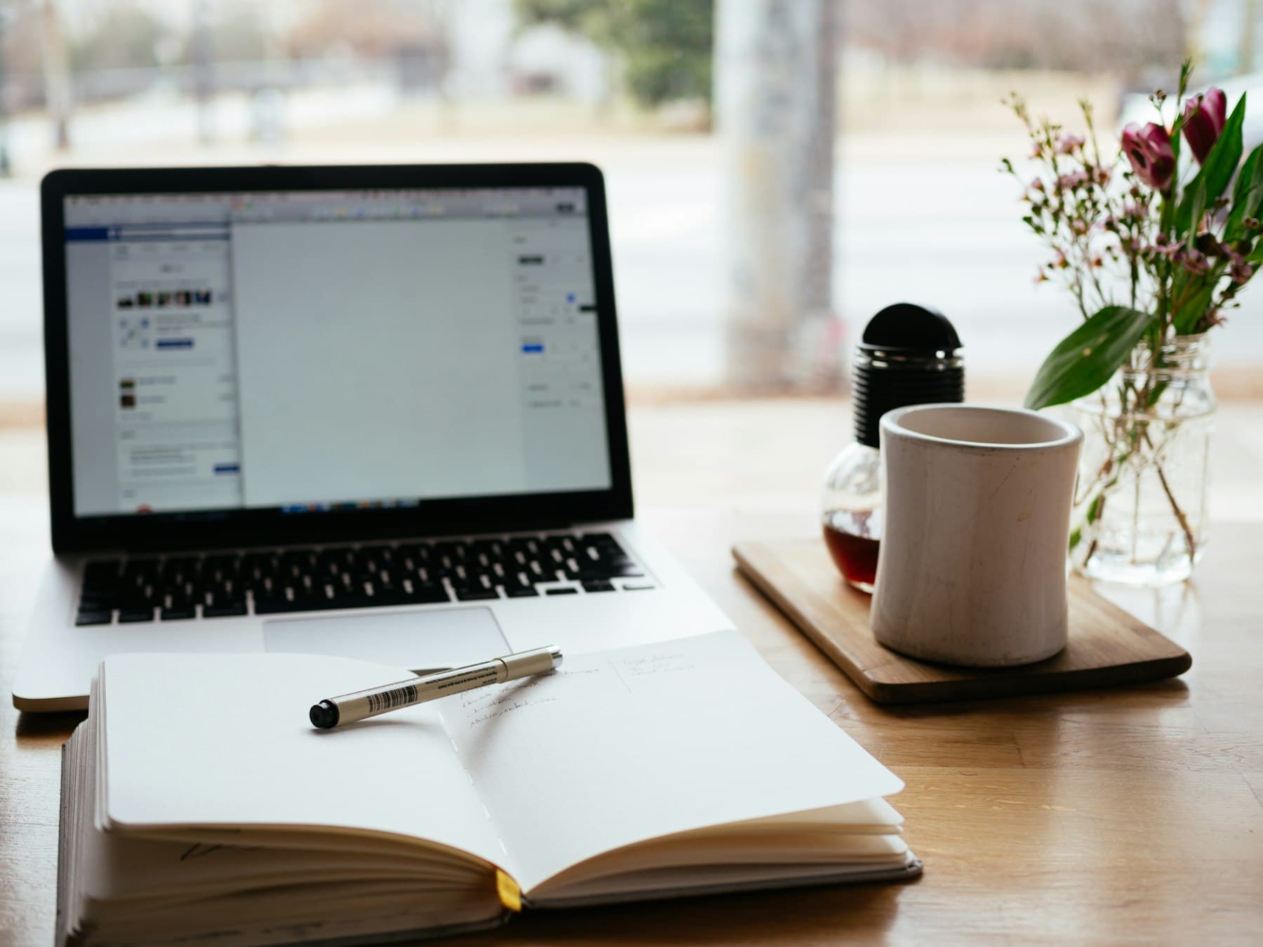 An open notebook and pen on a café table beside a laptop and a cup