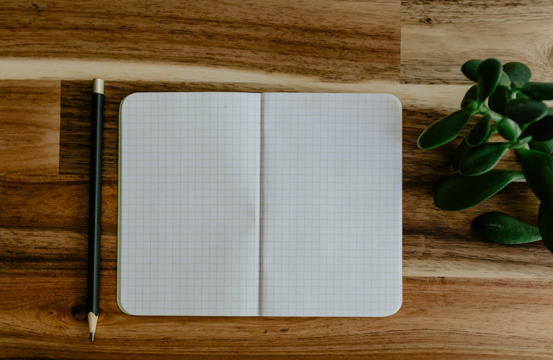 An open grid-ruled notebook lying on a wooden desk beside a pencil