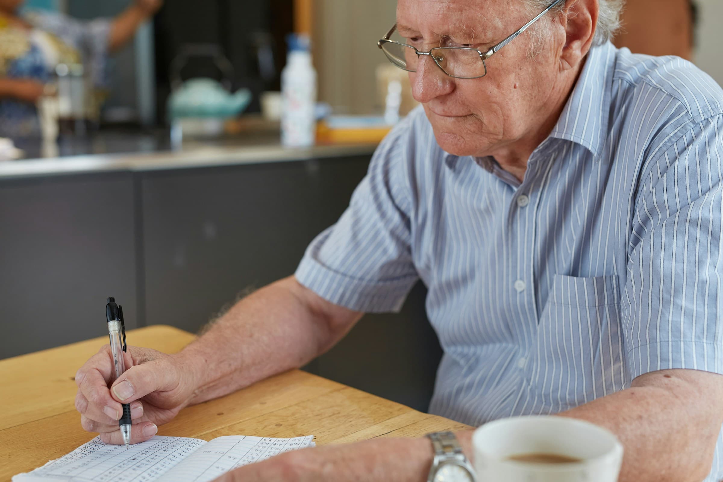 An older man writing with a pen at a table beside a cup of coffee